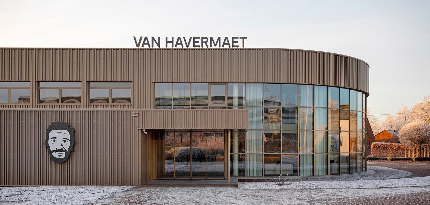 De moderne buitenkant van het Van Havermaet-kantoor in Westerlo. Een lichtbruine gevel met veel glas en een groot afgerond raampartij.